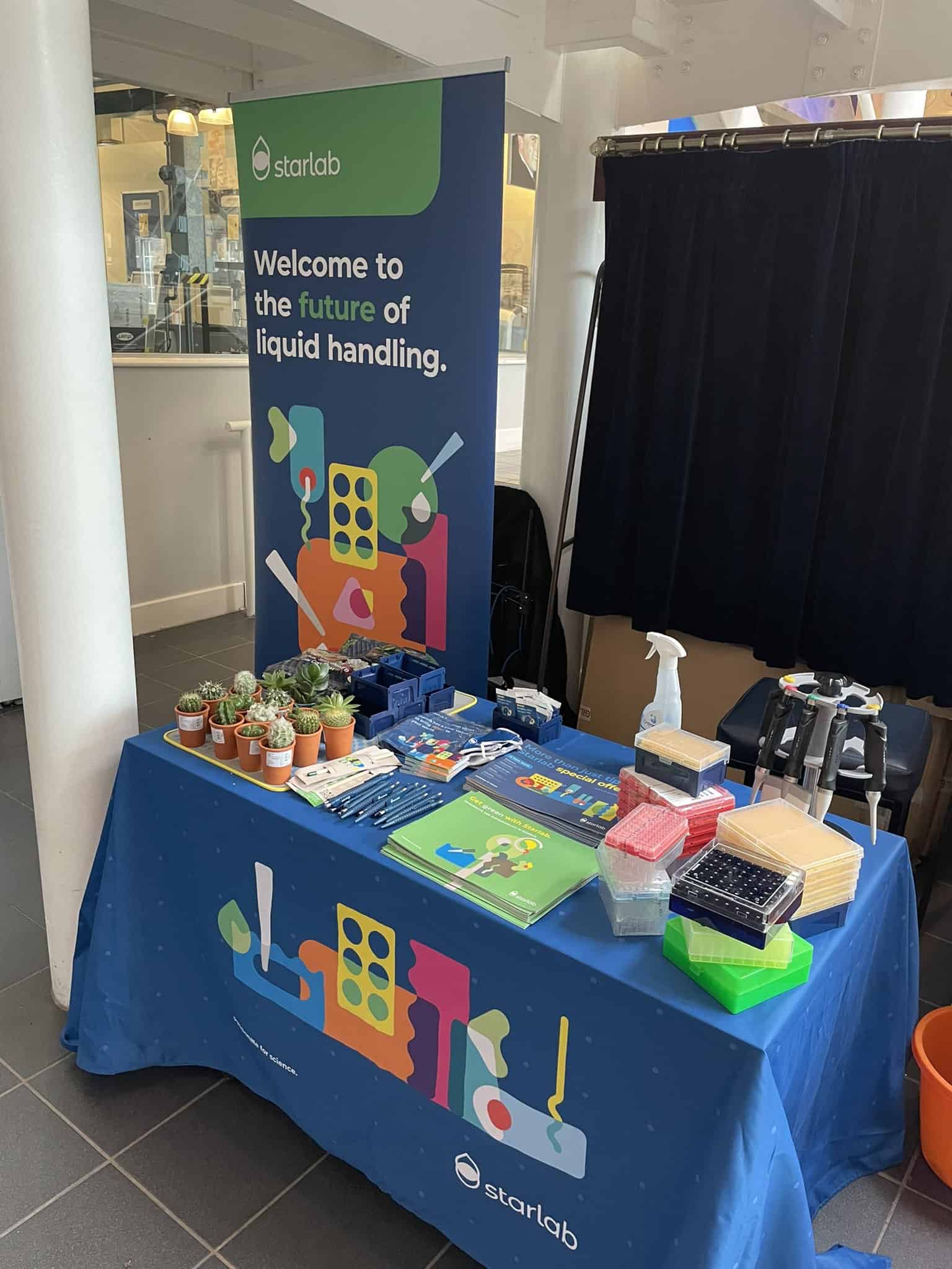 Image of STARLAB promotional stand. The table cloth is blue and the table is full of promotional material including pens, leaflets and cactus plants. 