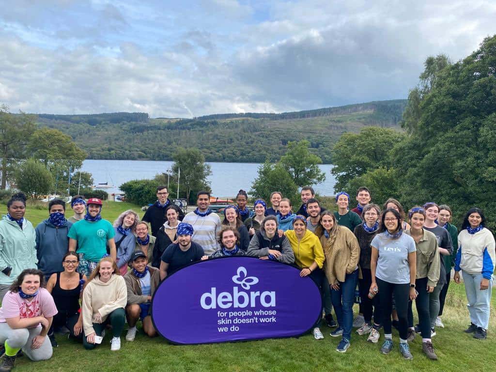 Photo of CDT students with a large purple DEBRA banner. The Coniston Lake is in the background