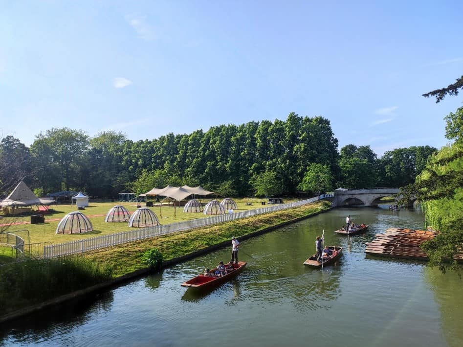 Photograph of a river in Cambridge. There are teepees and camping pods in the background, trees and grass with people paddling boats in the water.
