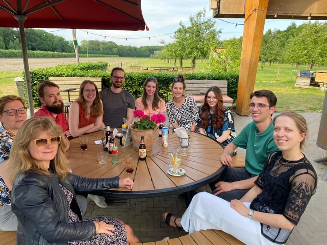 Group photo of Chloe with her placement colleagues. 10 people all sit around a round garden table with a red umbrella above them, there is grass, trees and fairy lights in the background and the weather looks warm and sunny.